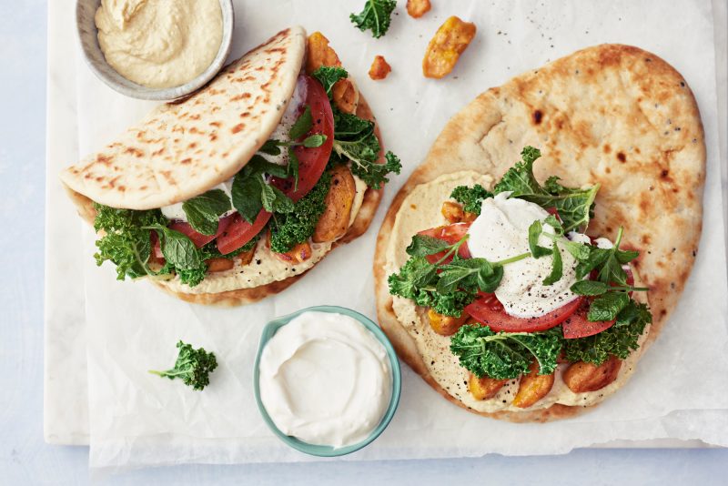 Kuvassa yksi naanleipä täytteiden kanssa näkyvillä. Täytteenä hummustahnaa, tomaattiviipaleita, yrttejä ja lehtikaalia. Kuvassa myös naanleipä, joka on jo taiteltu täytteiden kanssa ja hummus ja jogurttikastike omissa kipoissaan.