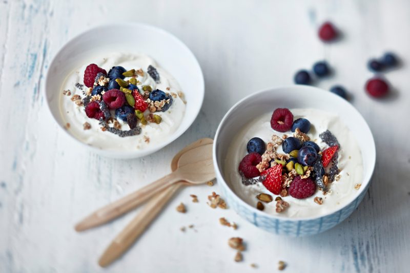 Maitojauheesta itse tehtyä jogurttia kahdessa kulhossa. pöytätasolla. Jogurtti on koristeltu marjoin, pähkinöin ja siemenin.